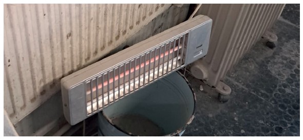 Ein Heizstrahler steht auf einem Eimer aus Eisen, direkt vor einem Heizkörper.