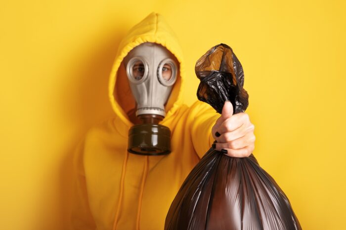 Person in gelbem Kapuzenpulli und Gasmaske, die einen schwarzen Müllsack vor einem leuchtend gelben Hintergrund hält. Konzept der Umweltverschmutzung oder Abfallwirtschaft.